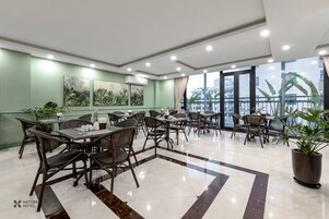 Un comedor de hotel con mesas y sillas, un ventanal con vistas, y una pared verde con obras de arte.