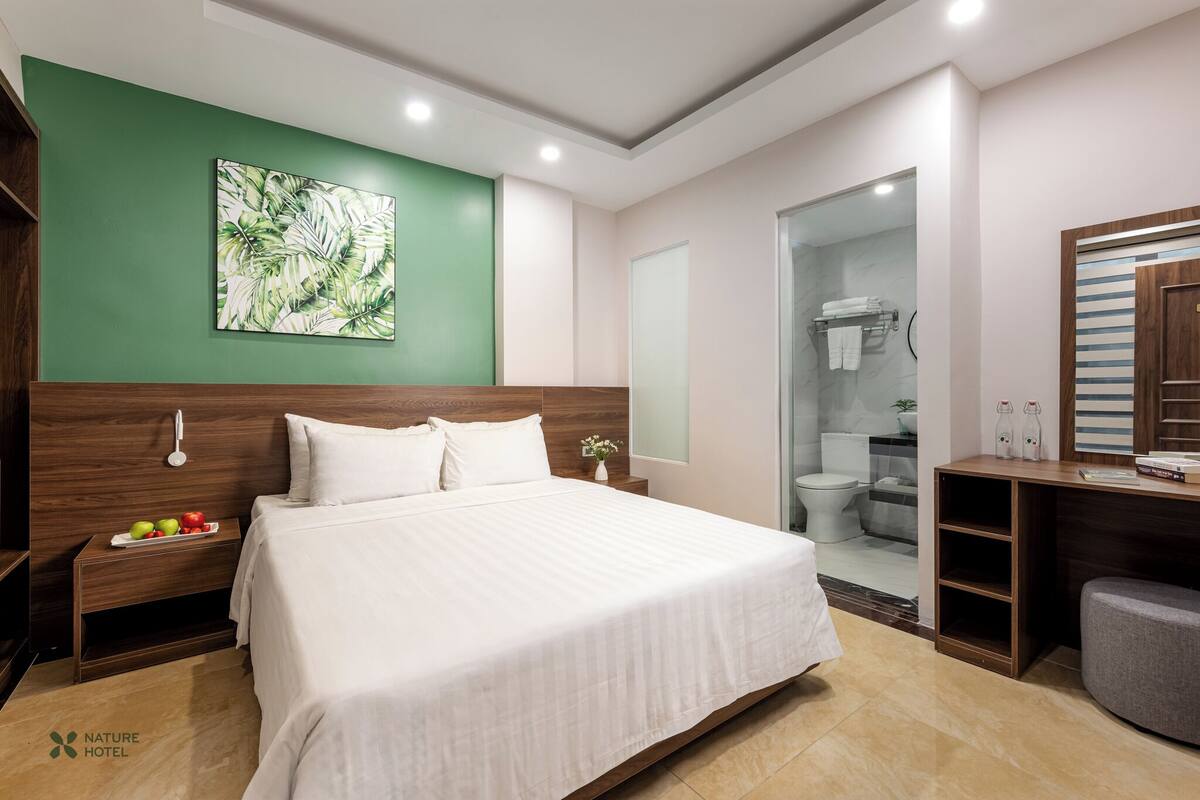 A hotel room with a large bed, a nightstand with fruit, a green accent wall, and a bathroom visible through a glass door.
