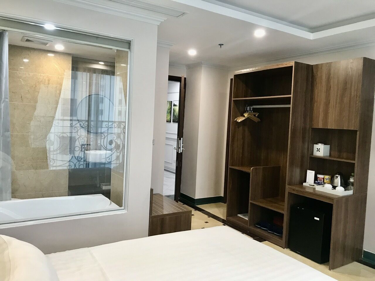 A hotel room with a large bed, a wardrobe with multiple shelves, a small desk, and a window with a view of the bathroom.