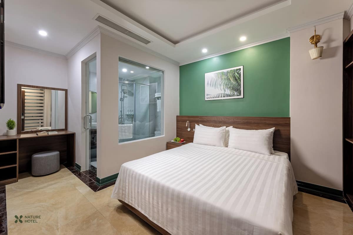 A hotel room with a large bed, a vanity with a mirror, a small stool, a nightstand with a lamp, and a green accent wall.