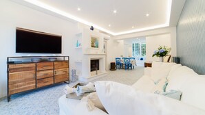 A modern living room with a flat-screen TV, a wooden cabinet, a white sofa, a fireplace, and a dining area with blue chairs.