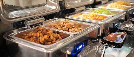 Un bufé con diversas opciones de comida en recipientes de acero inoxidable, una pantalla digital y una tetera.