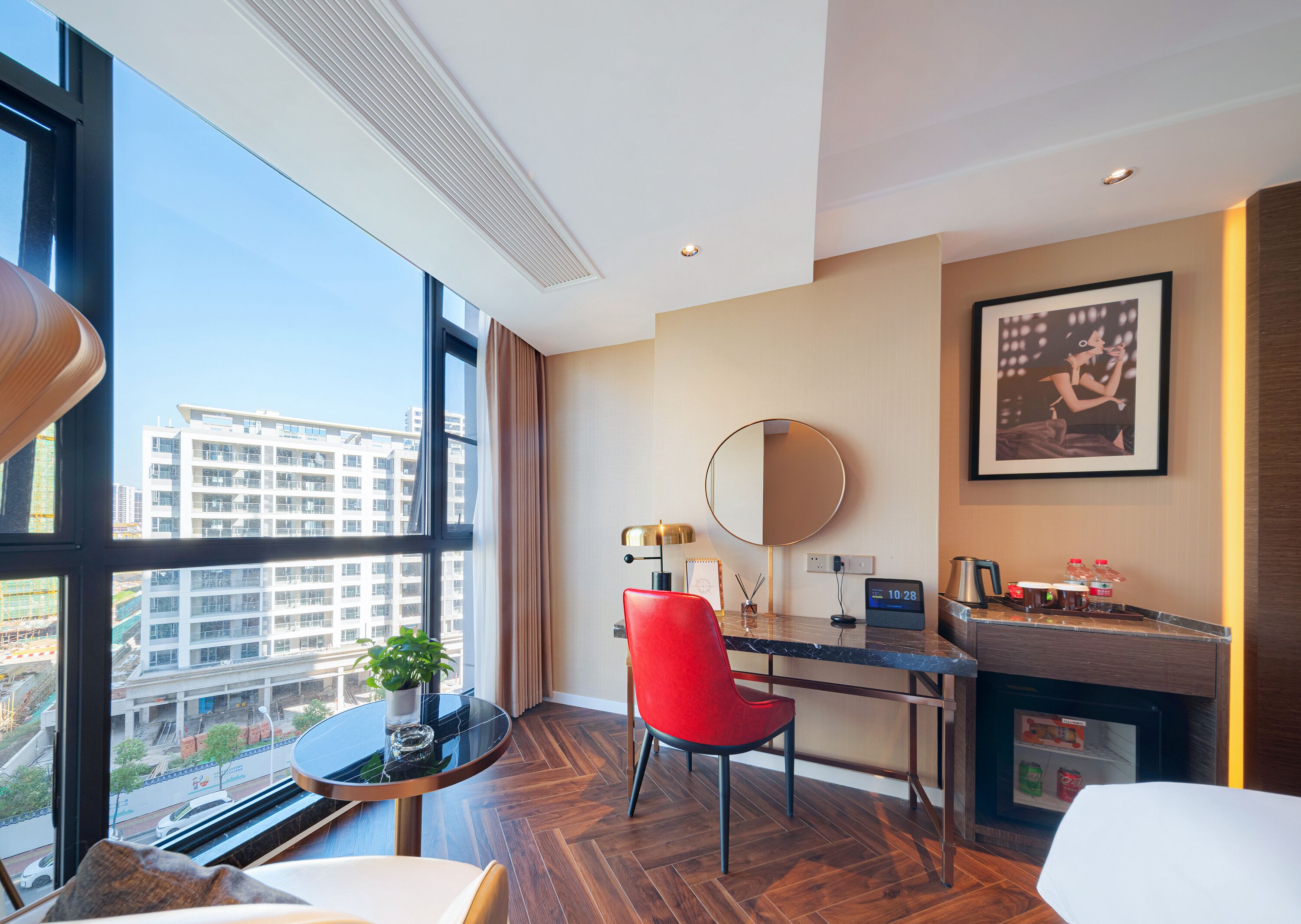 Habitación de hotel moderna con un amplio ventanal, un espejo redondo, un escritorio con una silla, una mesita con una planta y un minibar.