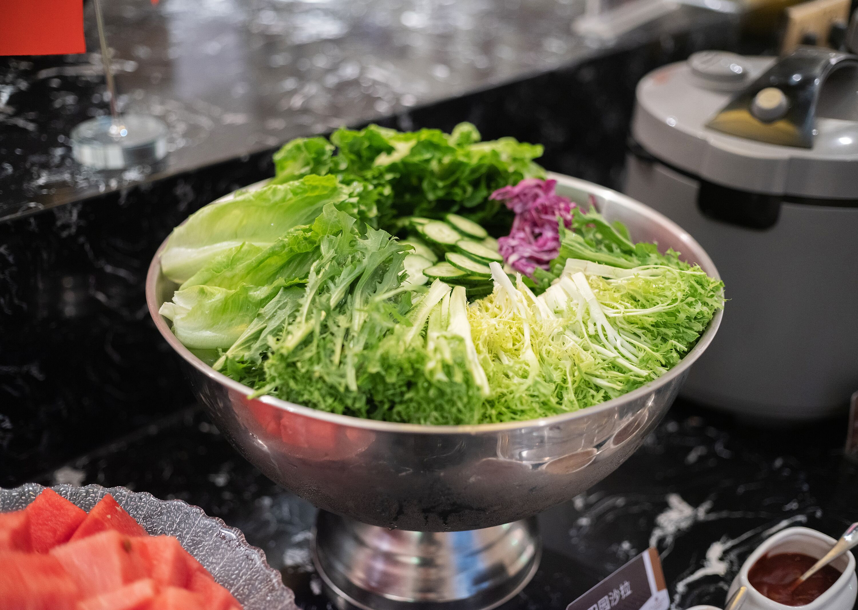 A metal bowl filled with various fresh vegetables and a small purple flower on a black countertop.