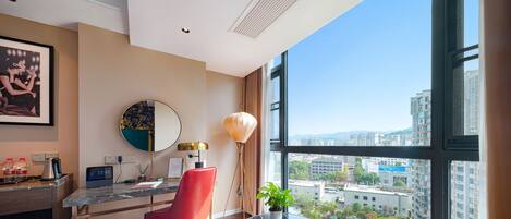Habitación de hotel moderna con un ventanal grande, un escritorio con computadora, una silla roja, una pequeña mesa con planta y vistas a la ciudad.