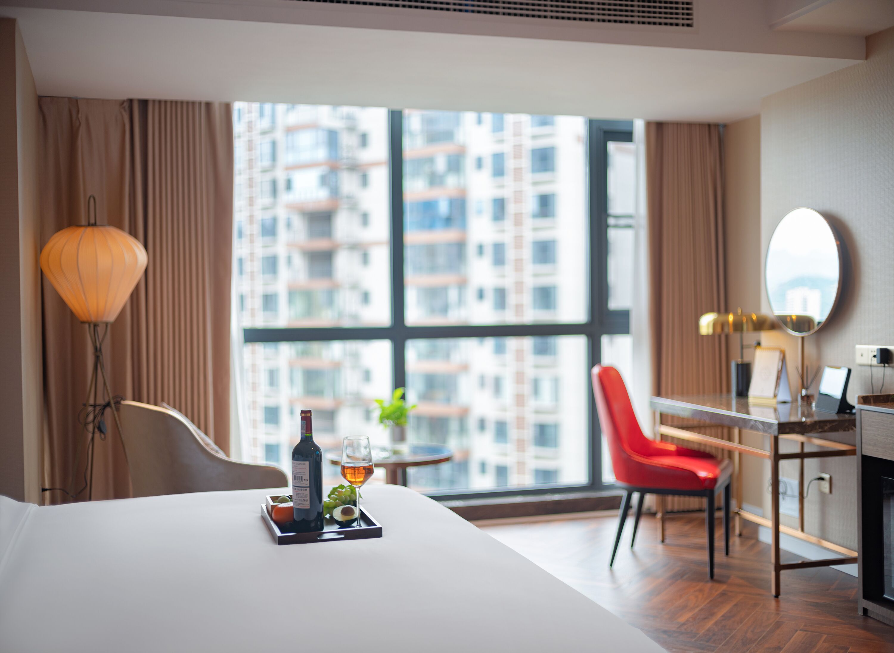 A hotel room with a bed, a desk, a chair, a window with a view of high-rise buildings, and a lamp.