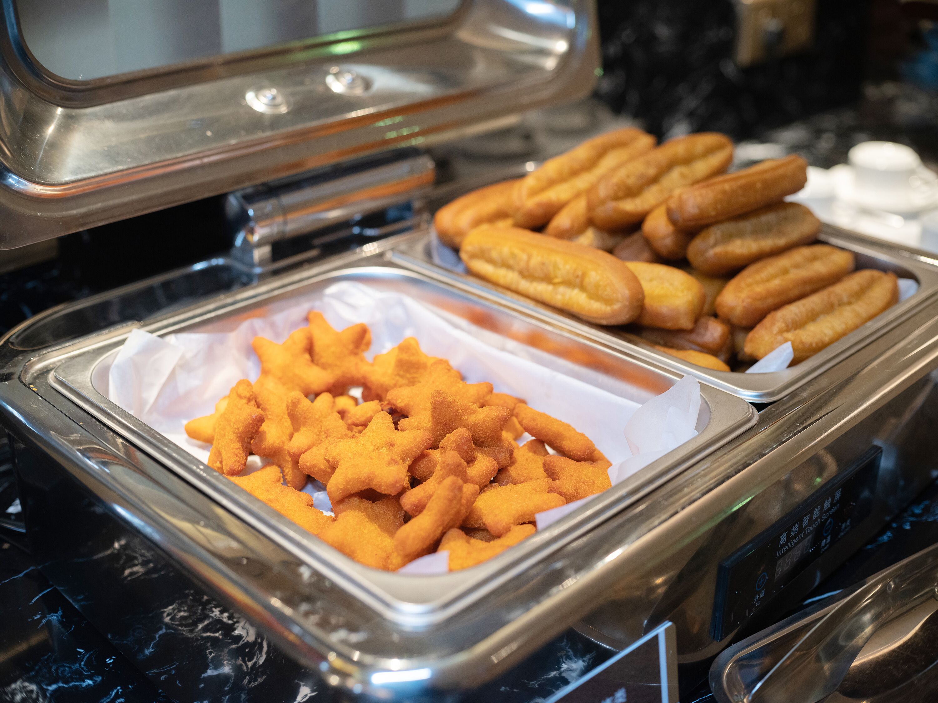 Un bufé con diversos alimentos fritos, incluyendo piezas con forma de estrella y otras cilíndricas largas, en una bandeja de metal.
