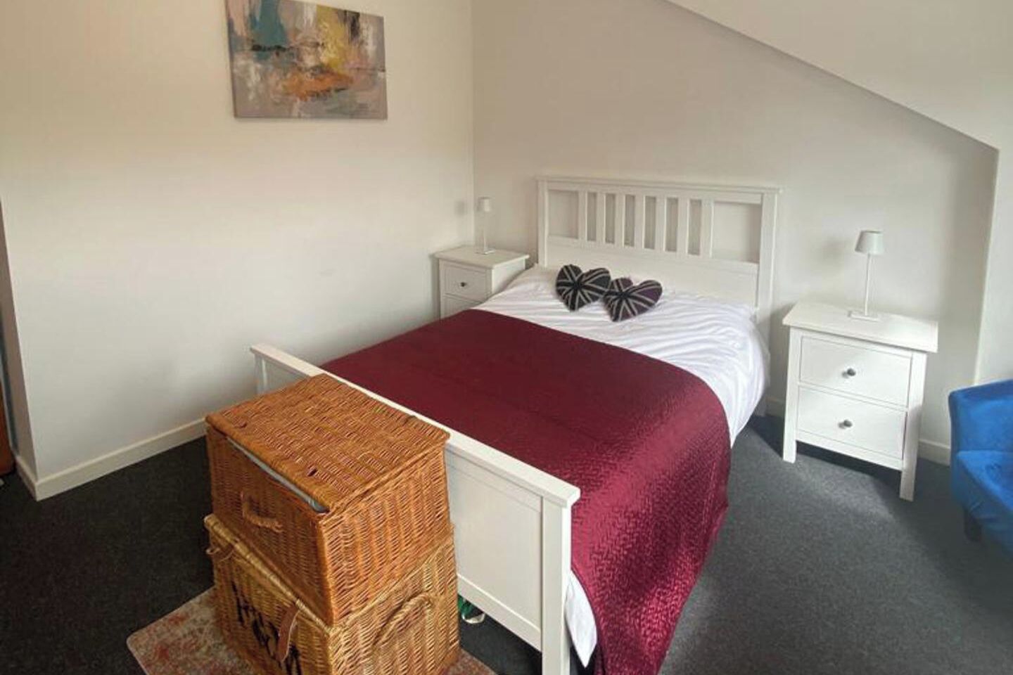 Makuuhuoneessa on sänky, valkoiset yöpöydät, punainen päiväpeite, sininen tuoli ja rottinkikori.