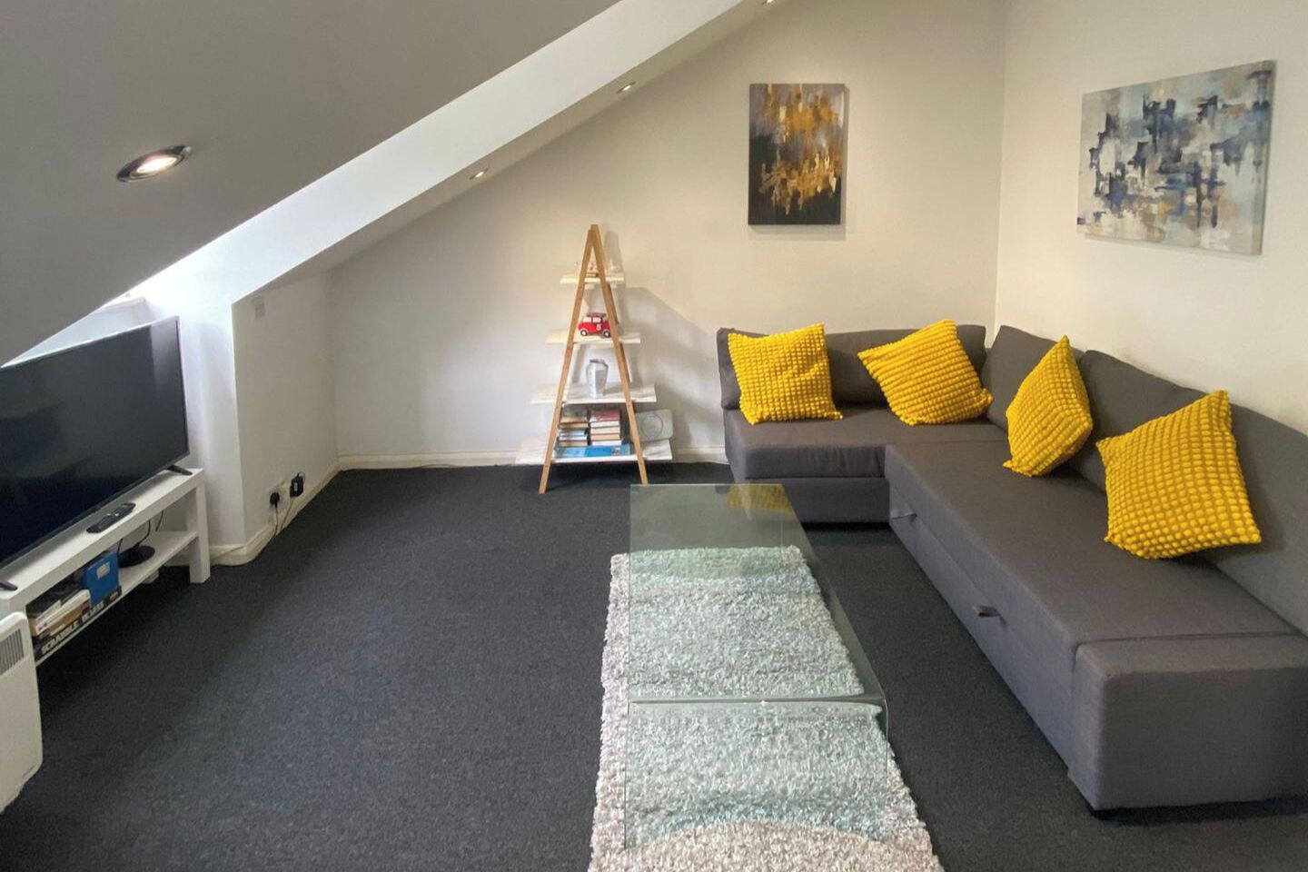 Ein modernes Wohnzimmer mit grauem Sofa, gelben Kissen, einem Fernseher auf einem Schrank, einem Bücherregal und einer an der Wand stehenden Leiter.