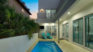 Un área de piscina moderna con una piscina azul cristalina, dos sillas y una puerta de cristal que da a un porche cubierto.