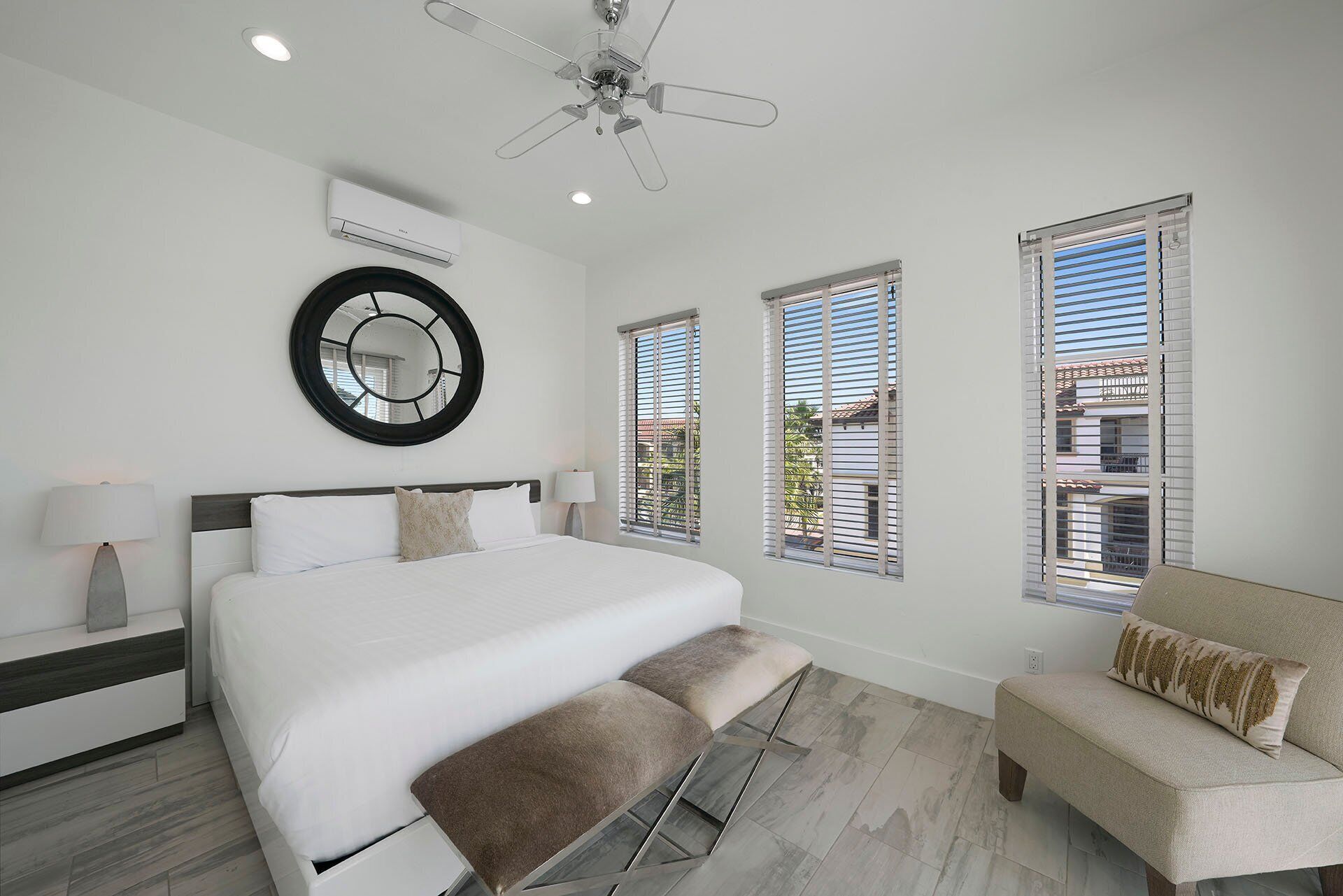 A modern bedroom with a large bed, a ceiling fan, and three windows with blinds.