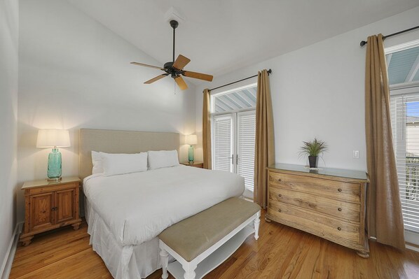 Een slaapkamer met een bed, een bankje, een dressoir, een plafondventilator en een raam met gordijnen.