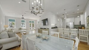 A spacious living area with a large white dining table, a sofa, and a kitchen area with white cabinets and a stainless steel refrigerator.