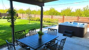 Un patio cubierto con una mesa y sillas de comedor, un jacuzzi y vistas a un césped y árboles bien cuidados.