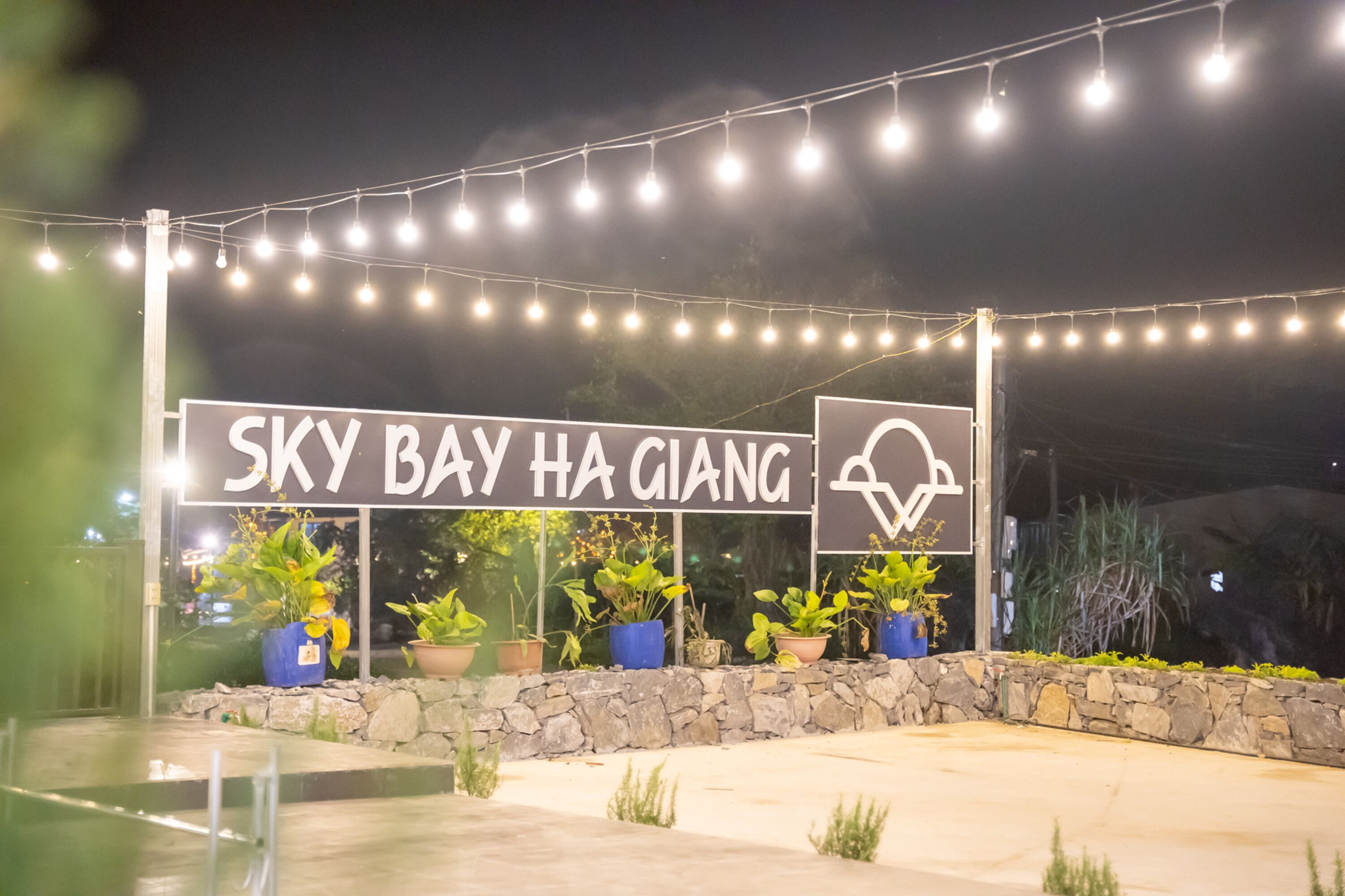 A sign for 'Sky Bay Ha Giang' with a mountain view symbol, surrounded by string lights and potted plants.