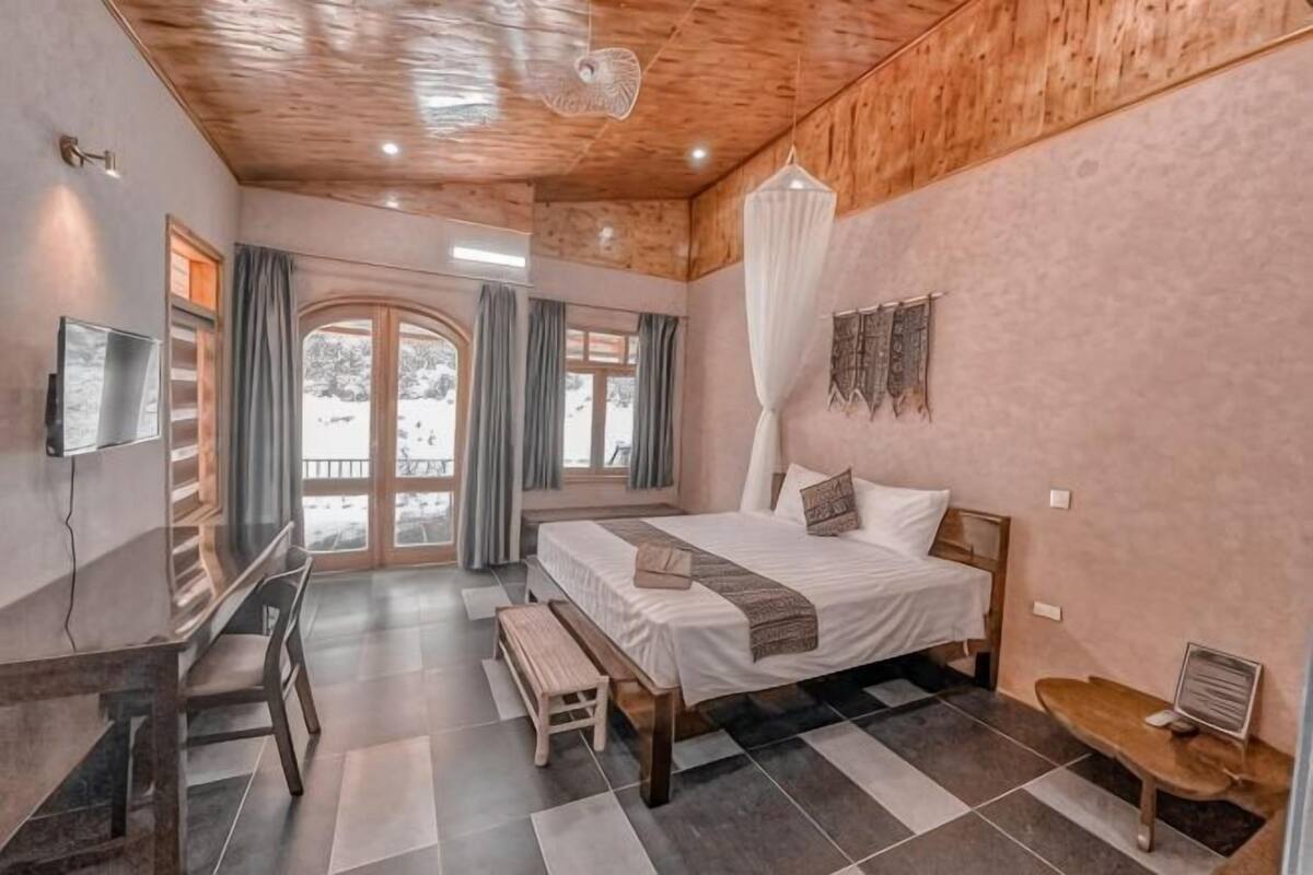 A bedroom with a bed, a desk, a chair, a bench, a TV, and a window with curtains.