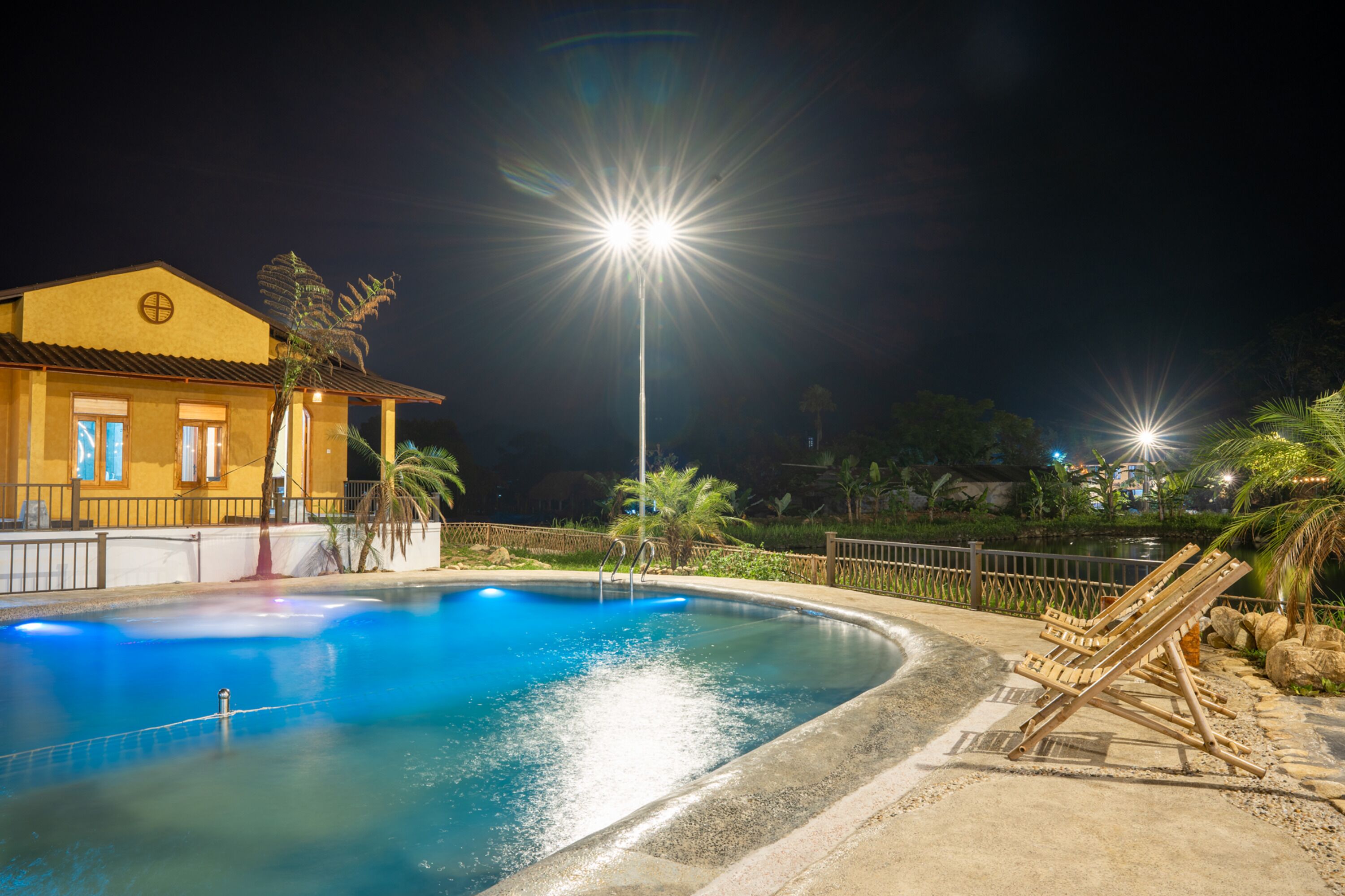 A nighttime swimming pool area with a house, palm trees, and a fence.