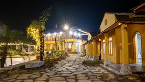 A stone pathway leading to a yellow building with a covered outdoor seating area and string lights. - Sky Bay Lodge (Ha Giang 1)