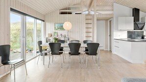 Una cocina moderna con armarios blancos y una campana extractora negra, un comedor con mesa blanca y sillas negras, y un amplio ventanal con vistas al exterior.