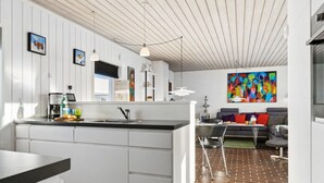 Cozinha moderna com armários brancos, bancada preta e piso de tijolo. Há uma área de jantar com sofá cinza e um quadro abstrato colorido na parede.