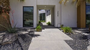 A modern building with a concrete pathway leading to a glass door entrance, surrounded by desert plants and rocks.
