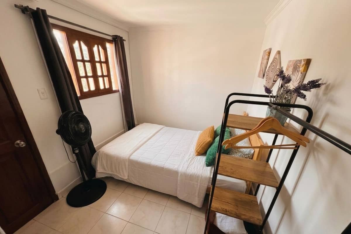 Un dormitorio pequeño con cama, ventilador, ventana con cortinas y un perchero de madera.