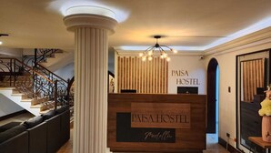 A reception area with a wooden desk labeled "PAISA HOSTEL" and a staircase with ornate railings.