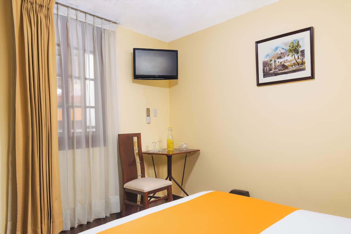 A hotel room with a bed, a television mounted on the wall, a small table with a chair, and a framed picture on the wall.