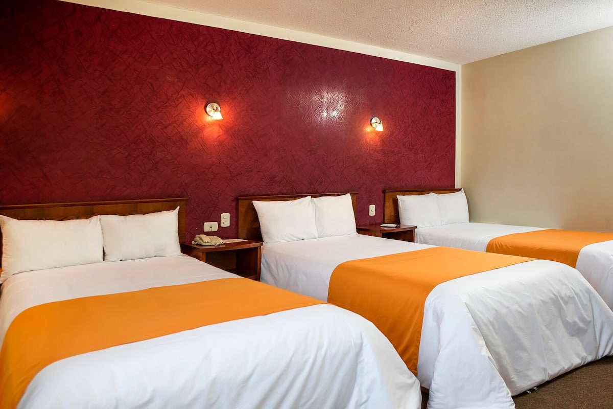 A hotel room with three beds, each with white bedding and orange accents. There are wall-mounted light fixtures and a wooden headboard.
