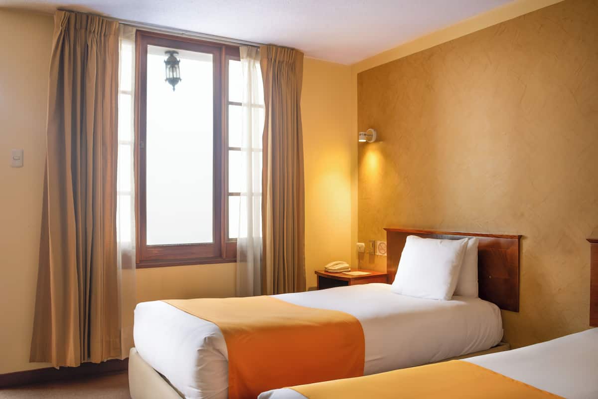 A hotel room with two beds, a window with curtains, a bedside table with a lamp, and a wall-mounted light.