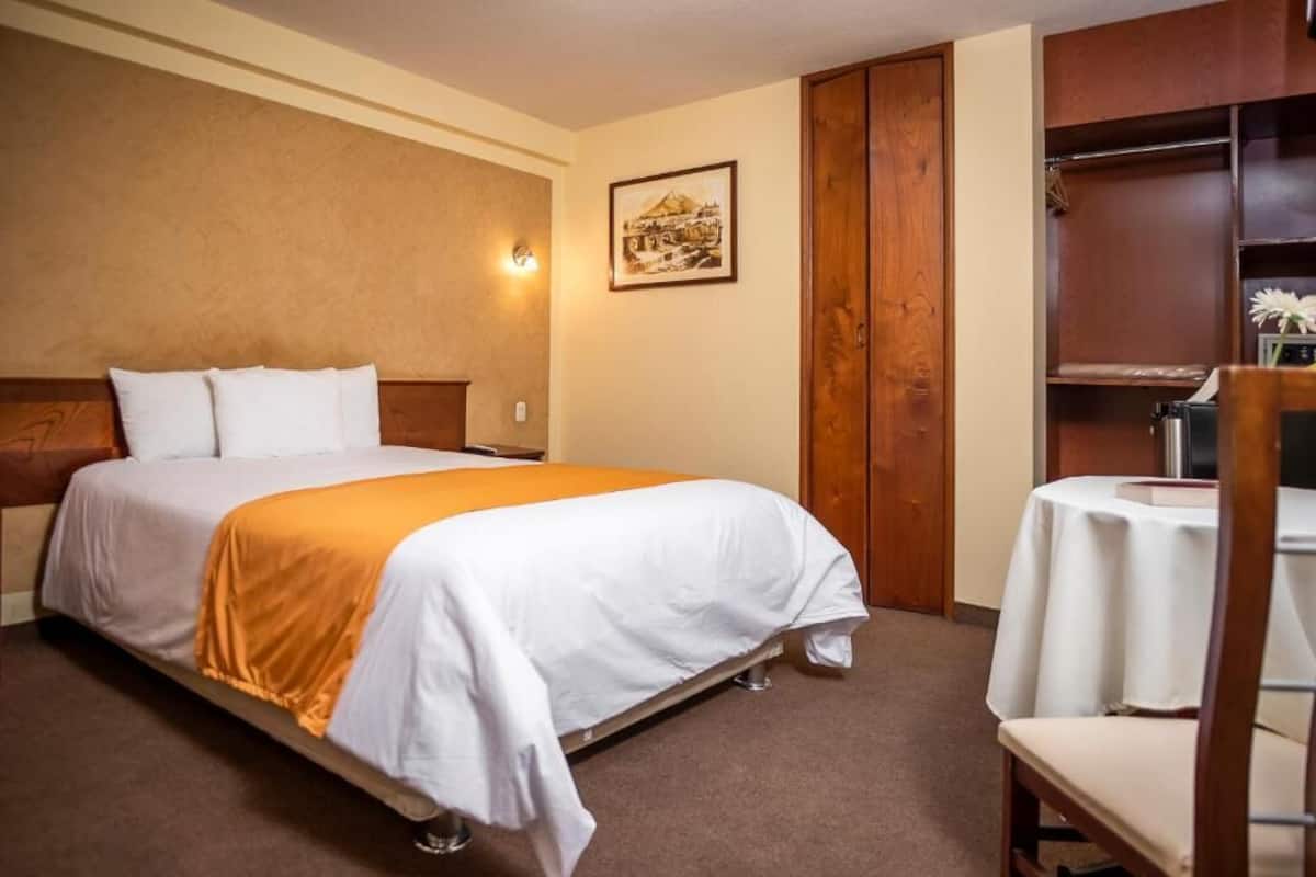 A hotel room with a bed, a wooden wardrobe, a small table with a chair, and a wall-mounted light fixture.