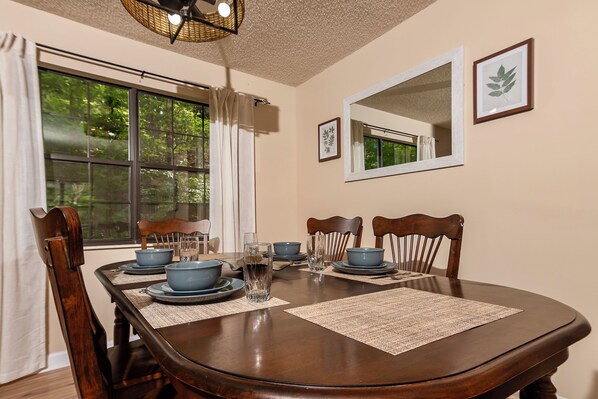 A dining room with a wooden table set for four, including blue bowls and plates, a pitcher of water, and a glass. There are two wooden chairs, a window with curtains, and a mirror on the wall.