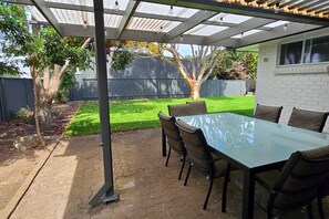 An outdoor patio with a glass-top table and chairs, surrounded by a brick floor and a metal pergola structure.