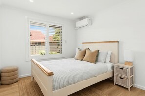 A bedroom with a bed, bedside tables, a window with blinds, and an air conditioning unit.