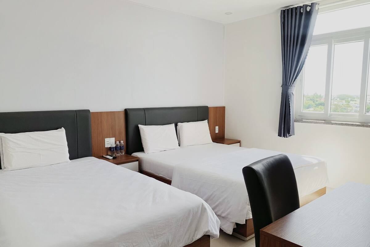 Een hotelkamer met twee eenpersoonsbedden, een nachtkastje van hout met een afstandsbediening en waterflesjes, een houten bureau en een raam met uitzicht op groen.