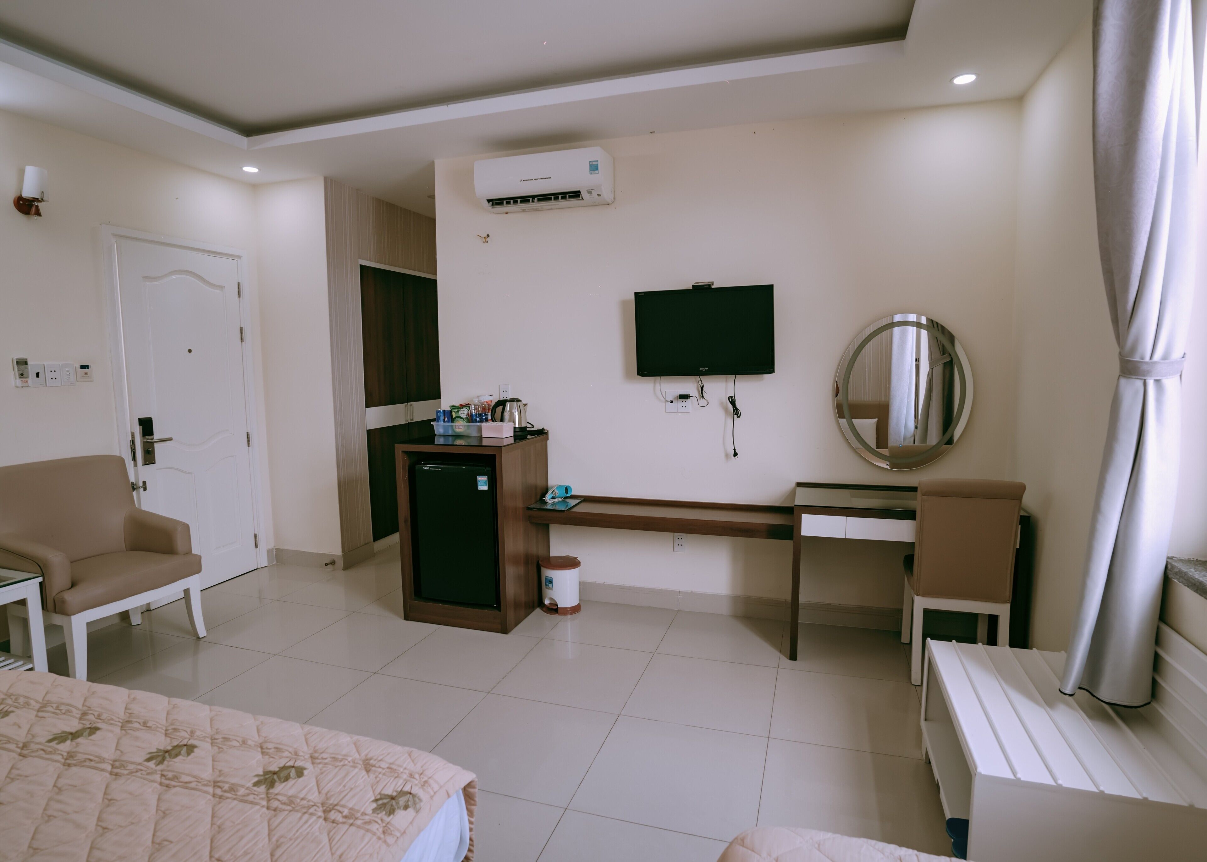 A hotel room with a bed, a desk, a chair, a small refrigerator, a television, and an air conditioning unit.