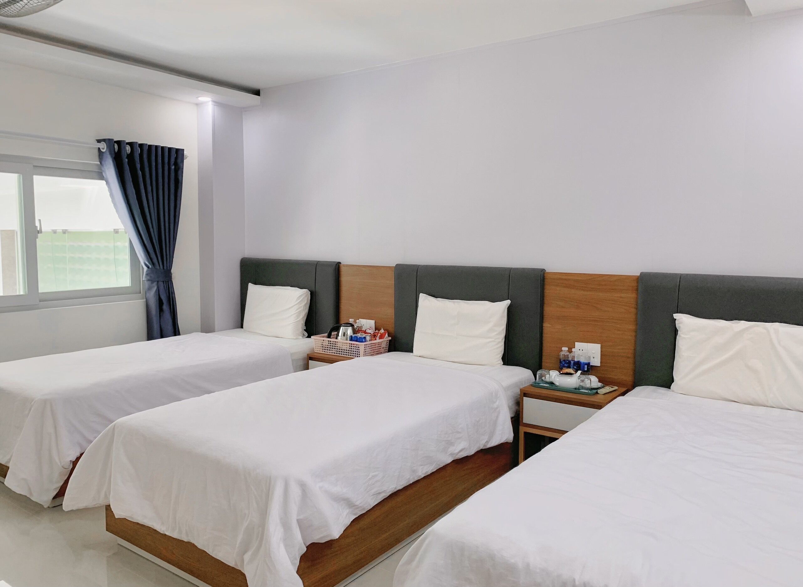 A hotel room with two single beds, a nightstand with a lamp, a small table with a tray of toiletries, and a window with curtains.