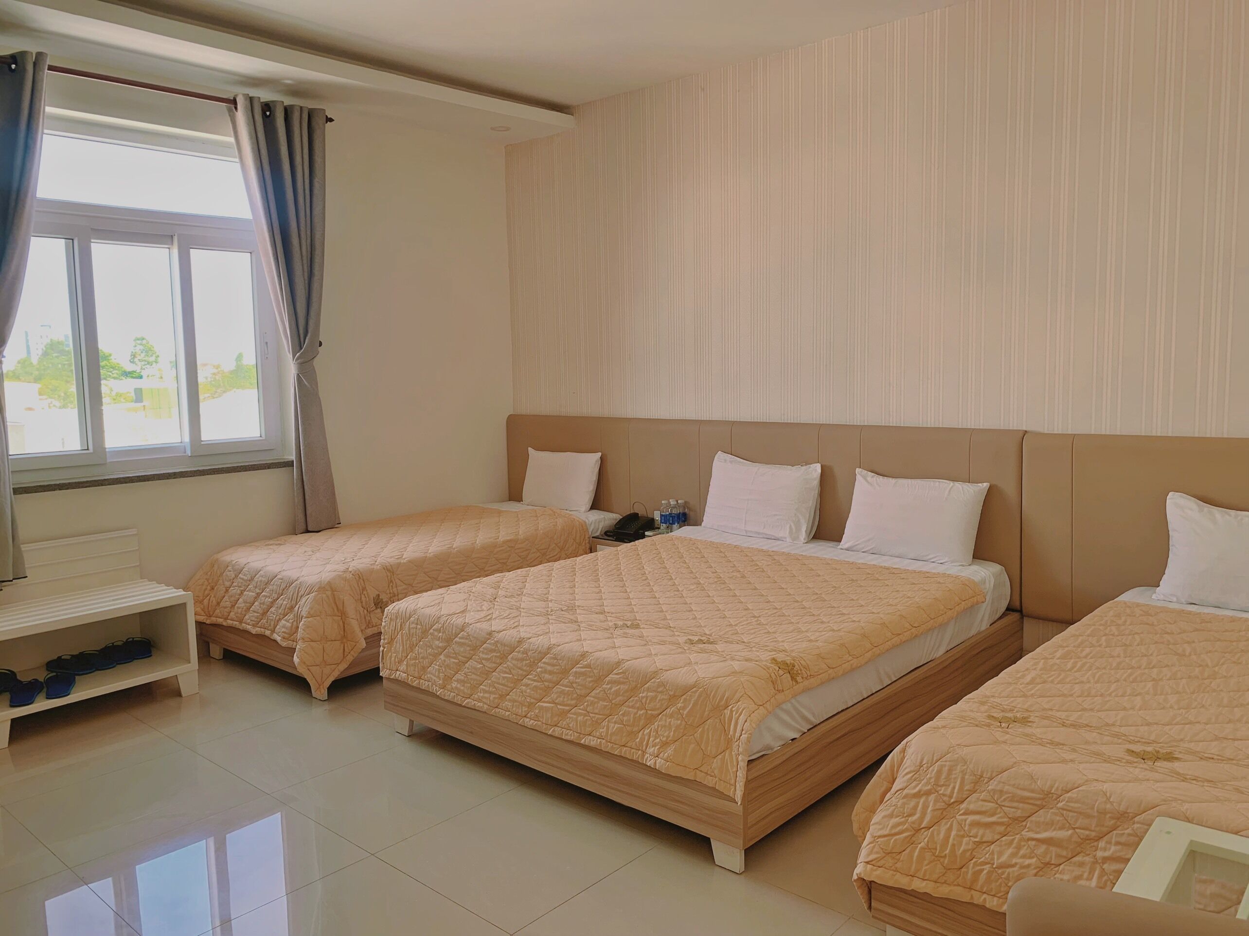 A hotel room with two beds, a bedside table, a shelf with shoes, and a window with curtains.