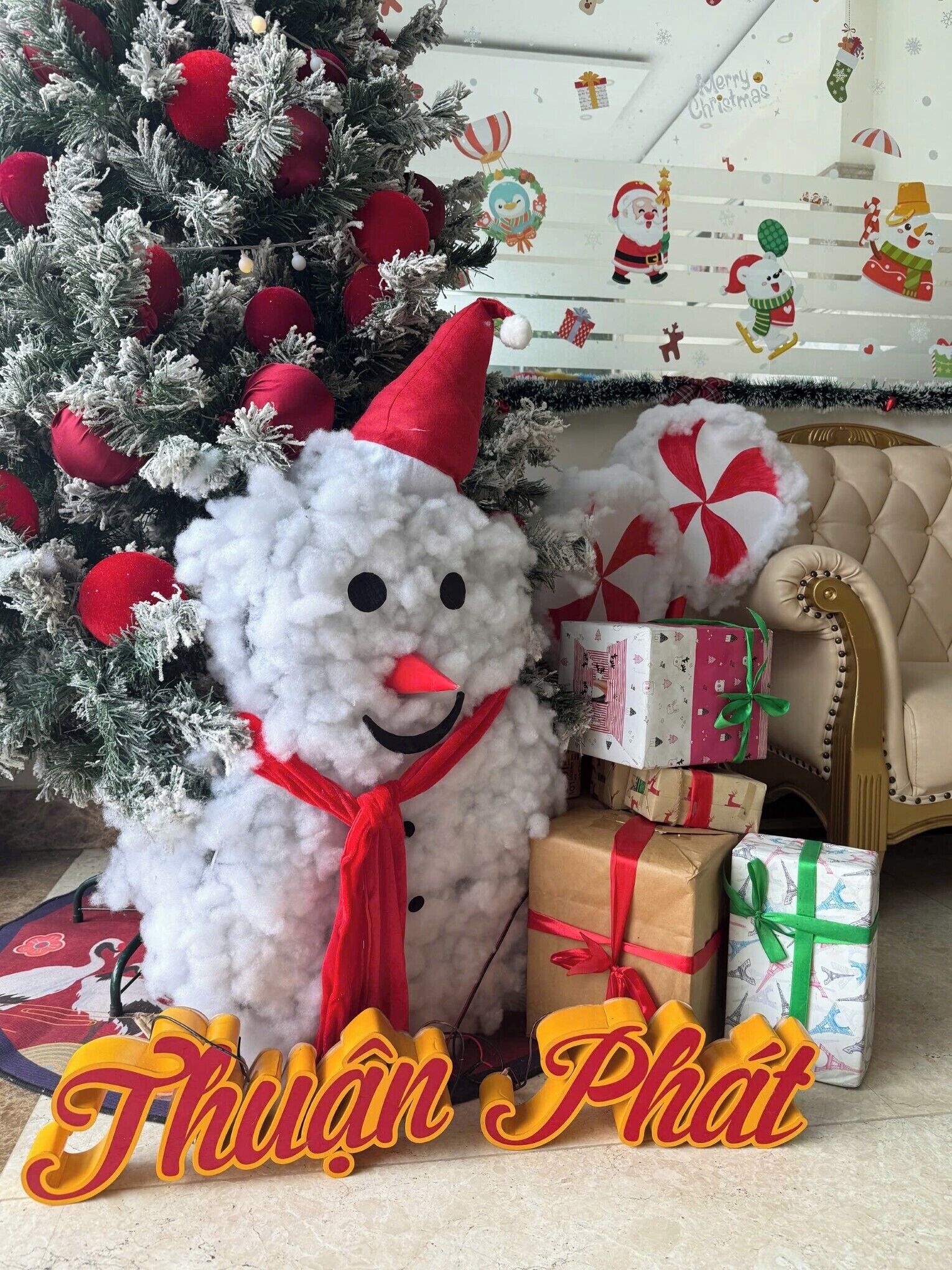 A snowman with a red hat and scarf, a Christmas tree with red ornaments, and wrapped gifts.