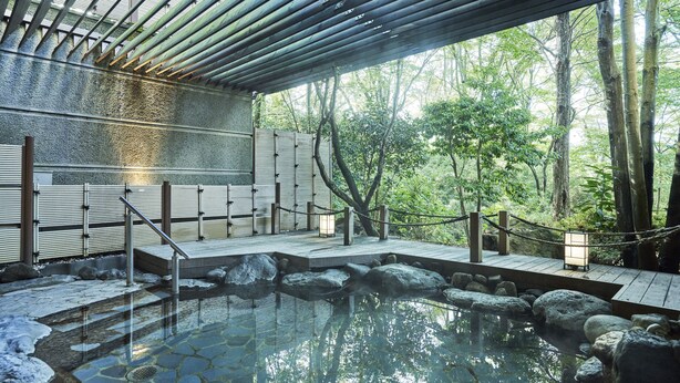 A traditional Japanese onsen with a wooden deck, stone steps, and a view of the surrounding forest.