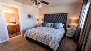 Ein Schlafzimmer mit einem gepolsterten Kopfteil, einem floralen Bettbezug, zwei Nachttischlampen, einem Deckenventilator und Blick auf ein Badezimmer mit Waschtisch und Handtuchhalter.