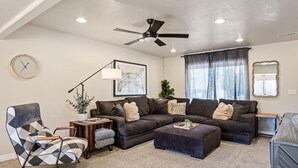 Ein Wohnzimmer mit einem Ecksofa, einem modernen Sessel, einem Couchtisch, einer Stehlampe, einem Deckenventilator und einem großen Fenster mit Vorhängen.