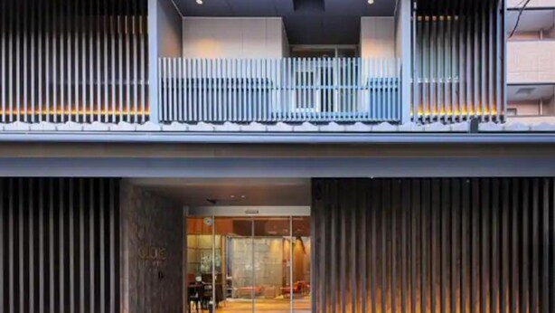 Un edificio moderno con entrada de vidrio, un balcón con persianas verticales y barandilla metálica.