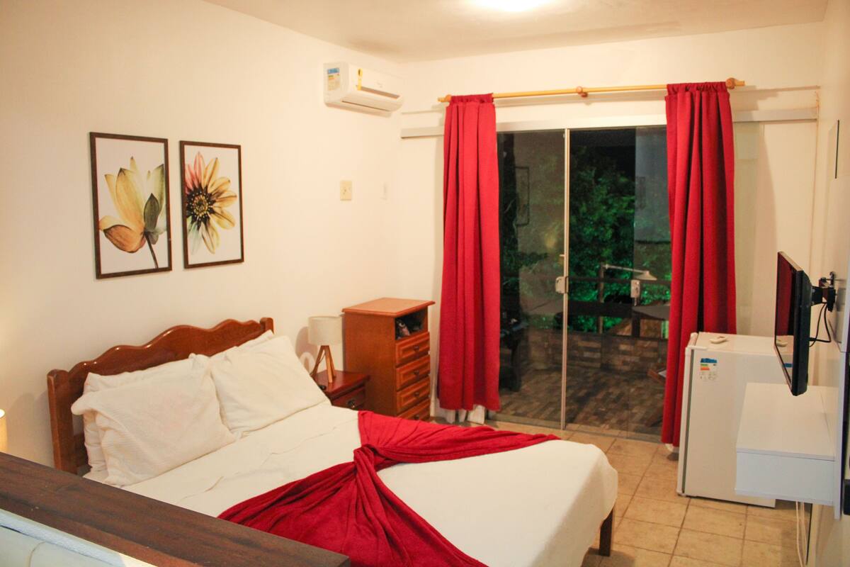 Una habitación de hotel con una cama, una mesita de noche de madera, un pequeño armario de madera, un refrigerador blanco y una puerta de vidrio corrediza con cortinas rojas.