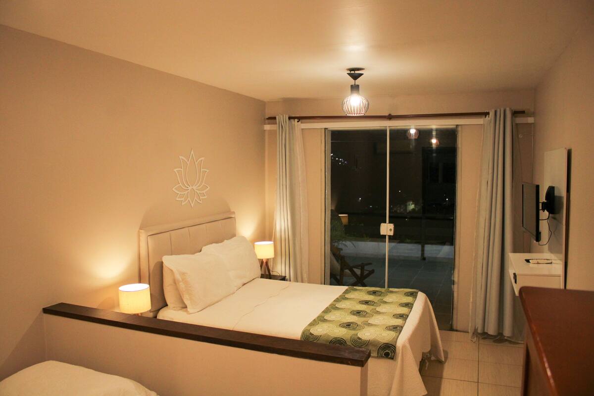 Habitación de hotel con cama, lámparas de noche, una mesa con mantel, televisión y puerta corrediza de vidrio que da a un espacio exterior.