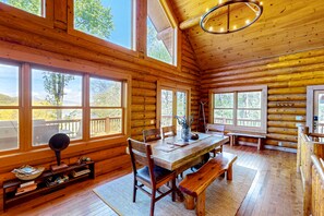 Cozy cabin interior with a high ceiling and large windows offering a view of the surrounding landscape. Features a dining table, chairs, and a bookshelf.