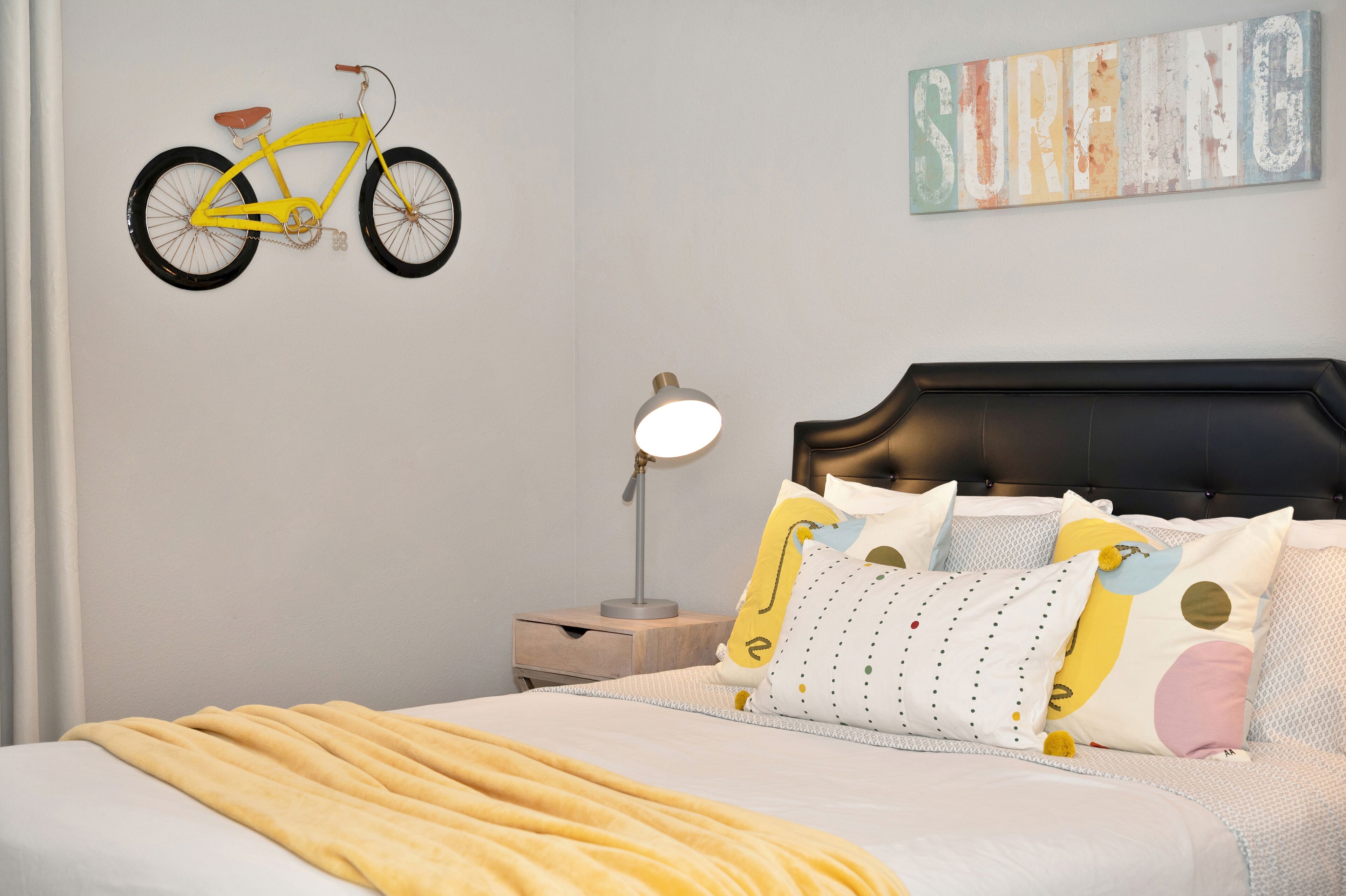 A bedroom with a bed, a nightstand, a lamp, a wall-mounted bicycle, and a surf-themed artwork.