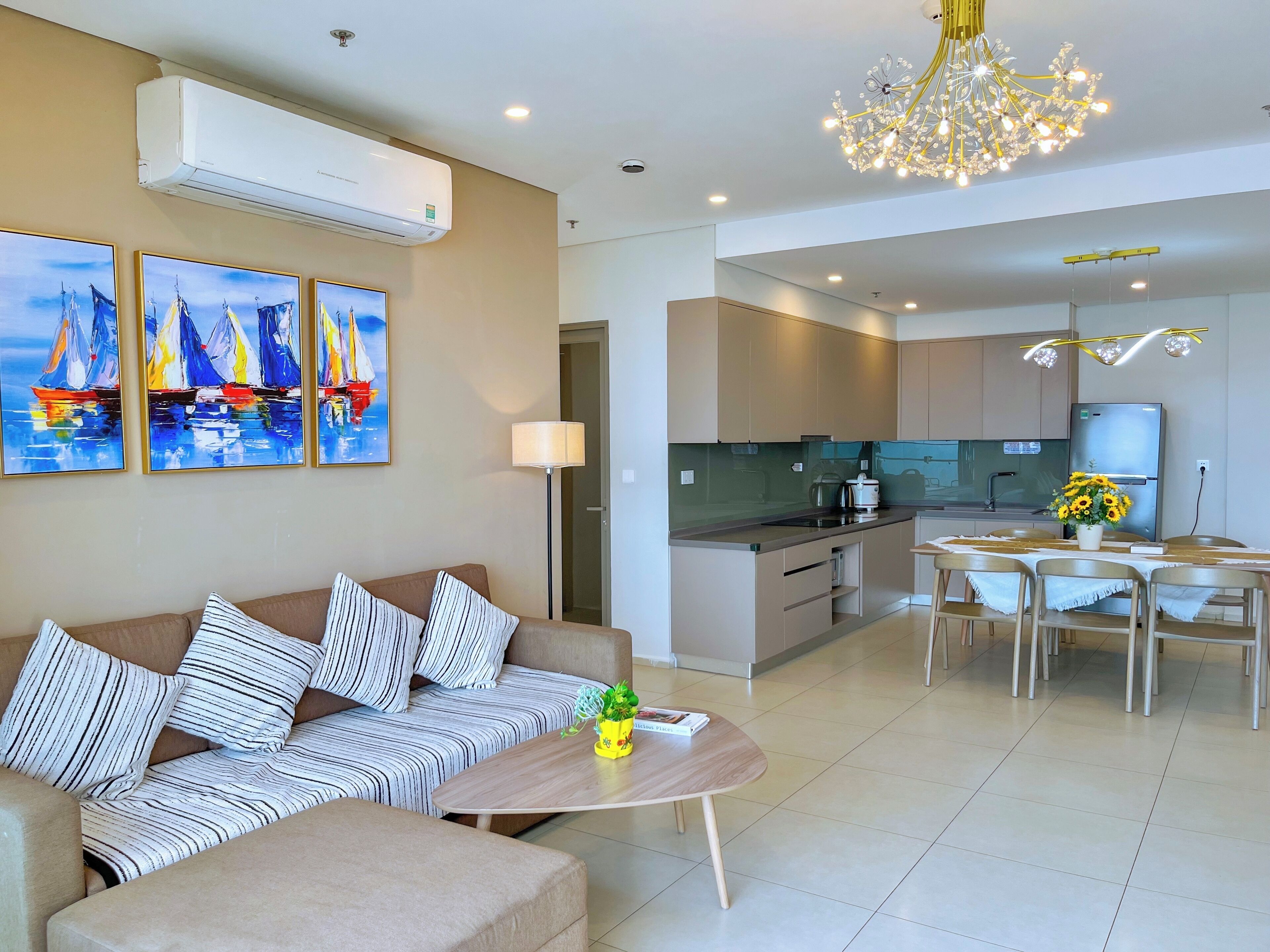 A modern living room with a sofa, a coffee table, and a dining area with chairs and a table. There is a kitchen in the background with cabinets and a refrigerator.
