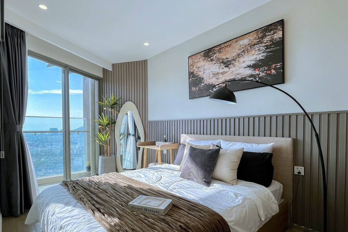 Habitación de hotel moderna con una cama grande, vistas de la ciudad, un cuadro en la pared y una lámpara de pie.