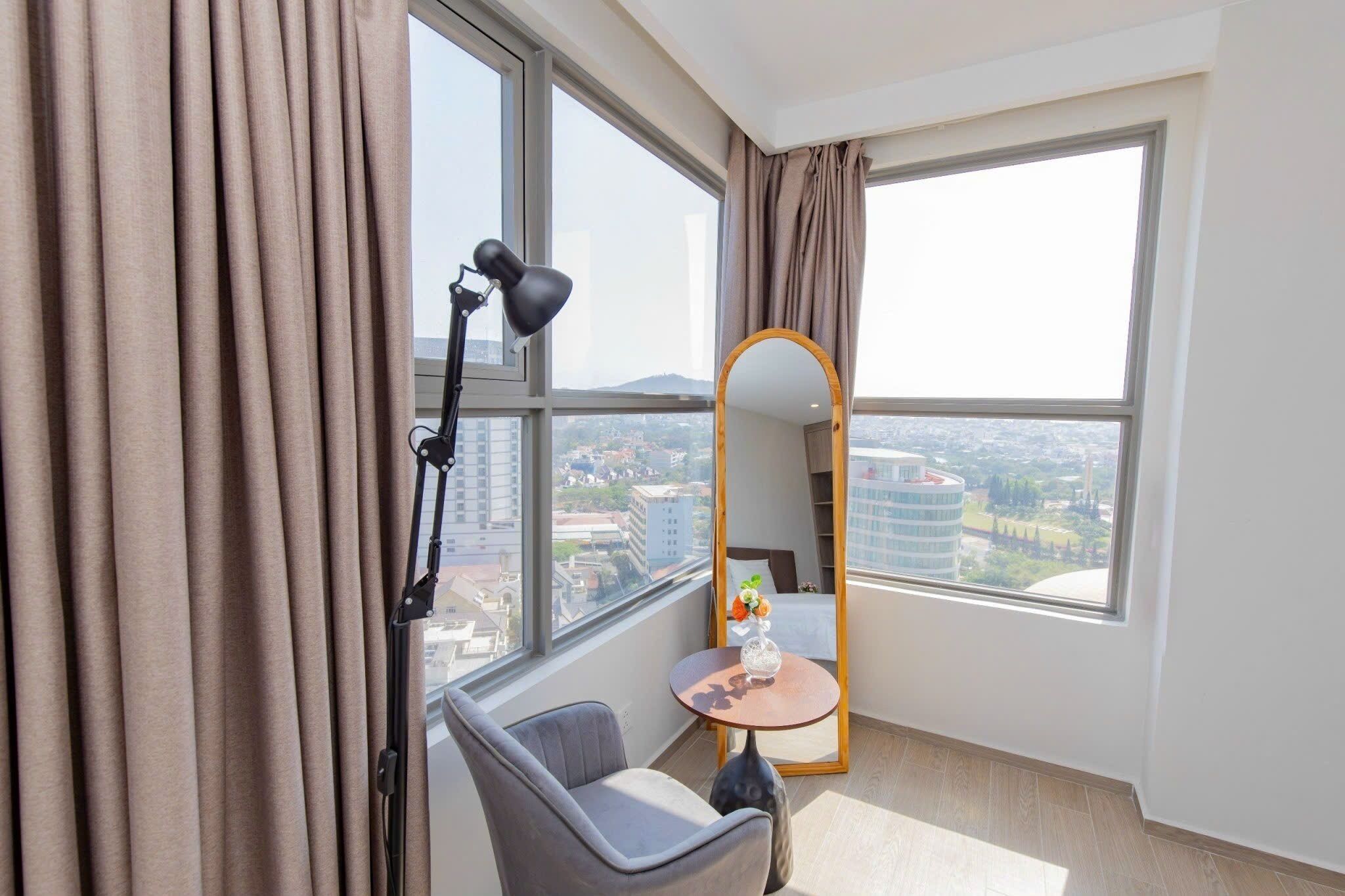 A modern hotel room with large windows, a round mirror, a desk with a vase, and a chair.